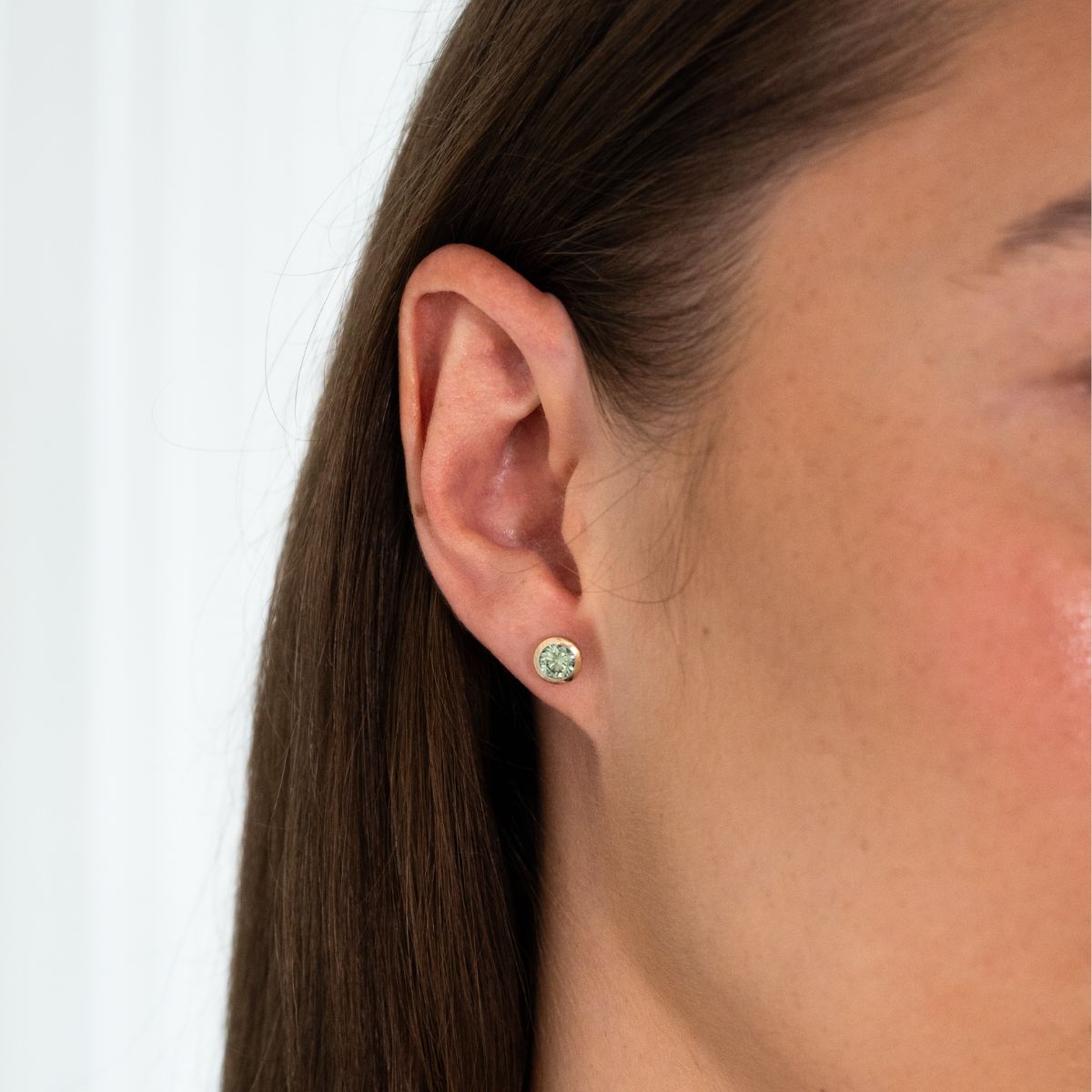 Nahaufnahme eines Ohrs mit einem kleinen, runden Ohrstecker mit grünem Diamanten in Zargenfassung in 0.5ct.