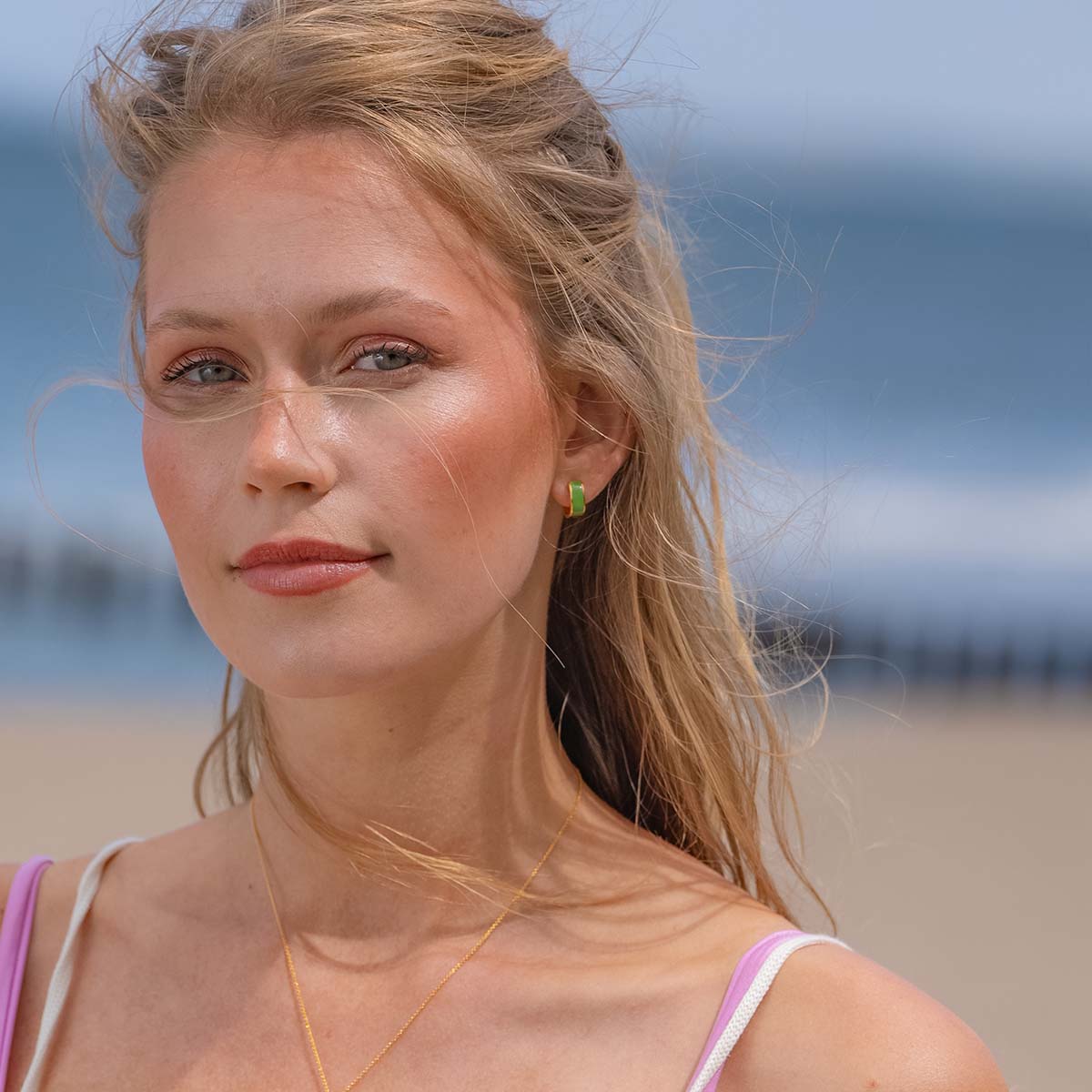 Model mit blonden Haaren blickt lächelnd in die Kamera und trägt grünen Ohrring am Ohr